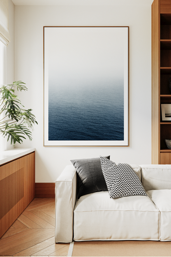 Modern living room with a white sofa, wooden bookshelf, and framed artwork of a blue ocean.