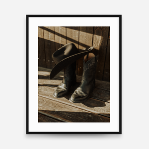 A framed art print featuring a photograph of black leather cowboy boots and a matching cowboy hat resting on weathered wooden planks, with dramatic diagonal shadows. The print is matted with a white border and enclosed in a slim black frame, set against a light gray background.