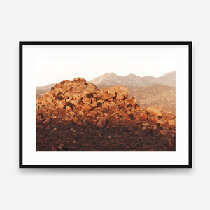 Fine art photograph of a Joshua Tree desert landscape at sunset with golden red light on rocky terrain and distant mountains, framed wall art by Lloyd Print Studio.